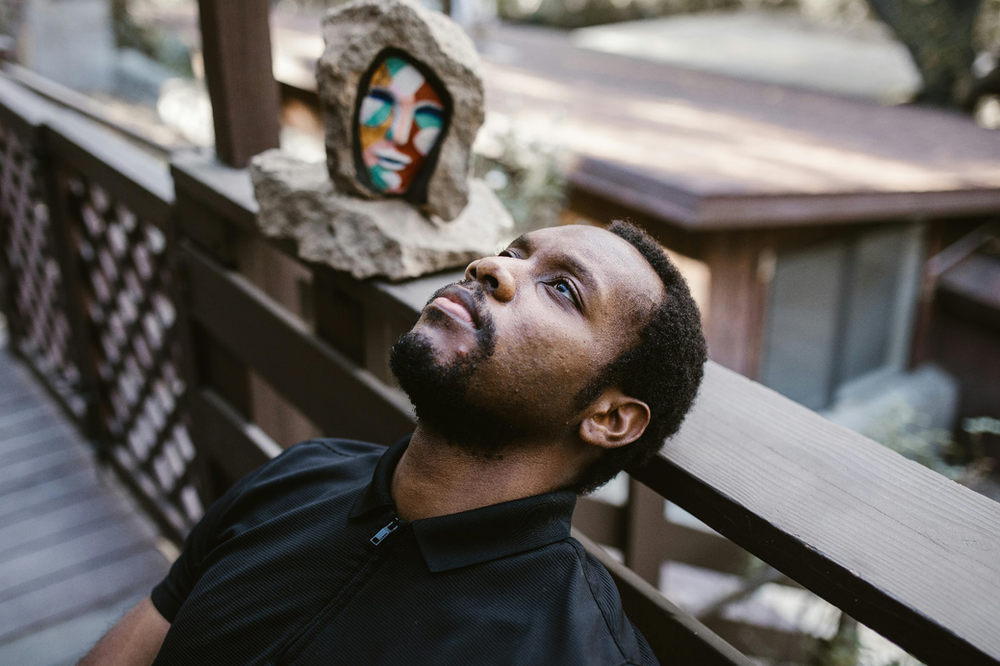 A man leans back on a railing outdoors in a black polo next to a rock with an abstract painting of a colorful face