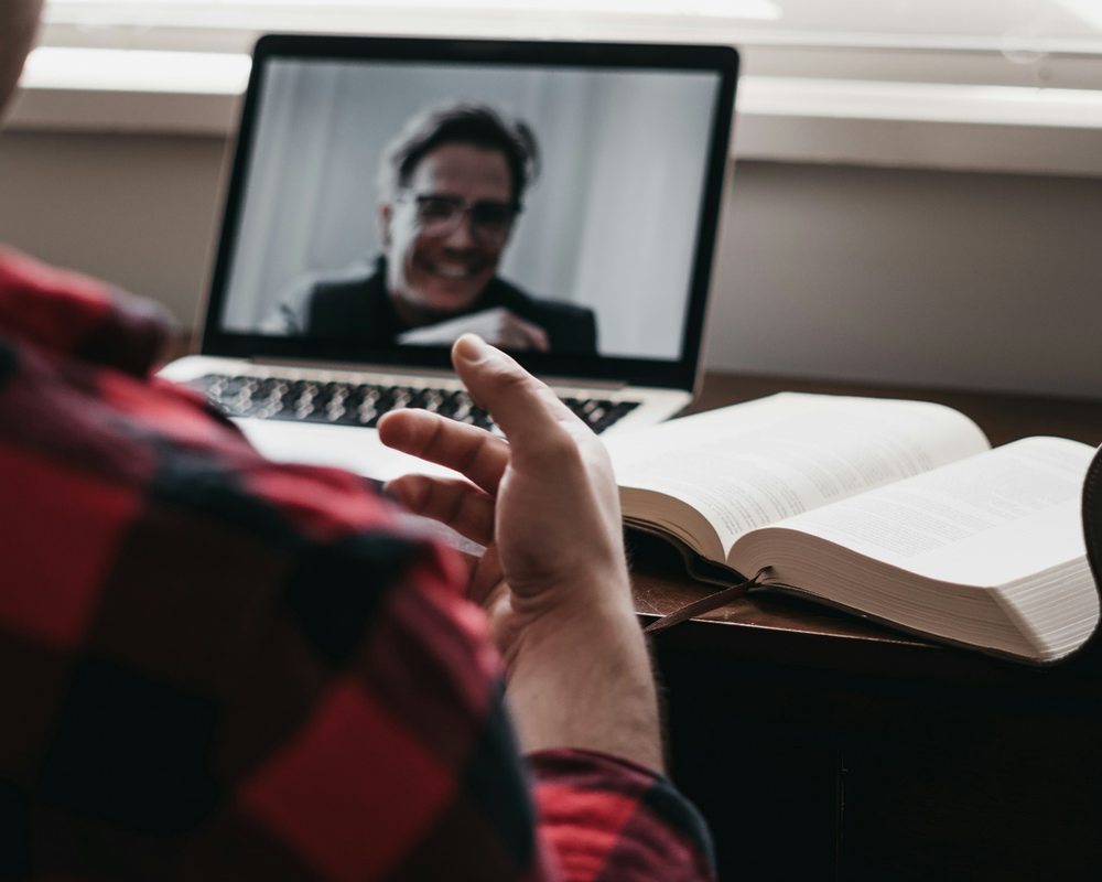 A person on a laptop screen opposite another with a book open on a desk showcasing what an online DBT program session may look like