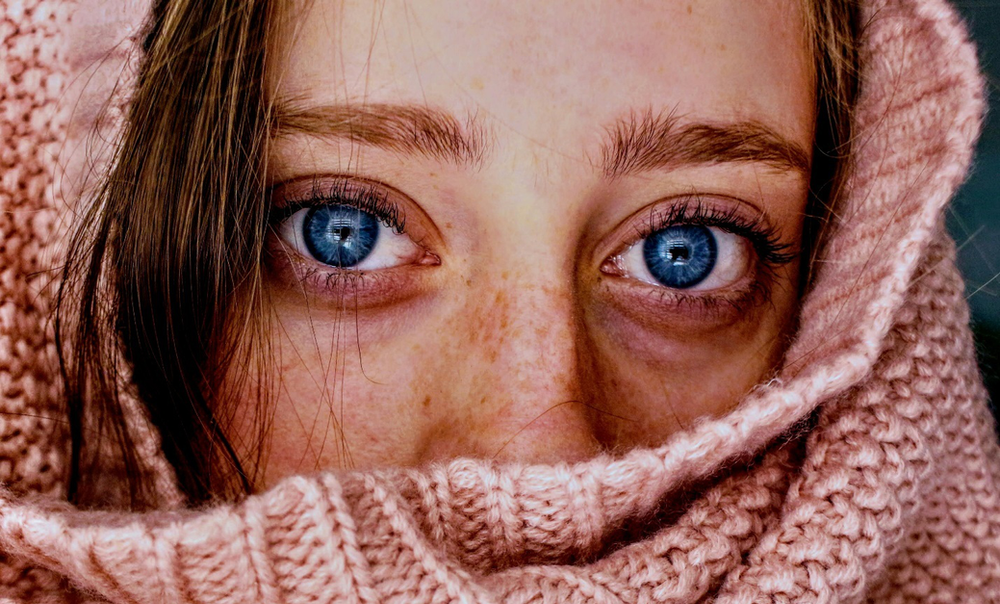 A close-up of a woman’s freckled face with a pink knitted textile hiding her head, tip of the nose and mouth