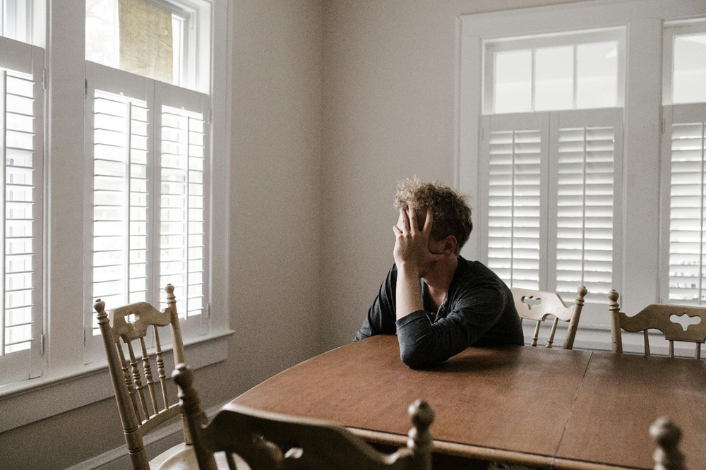 A person holds their head in their hand, resting their elbow on a dining table surrounded by chairs and white shuttered windows while looking out, symbolizing the need for more awareness around mental health for men